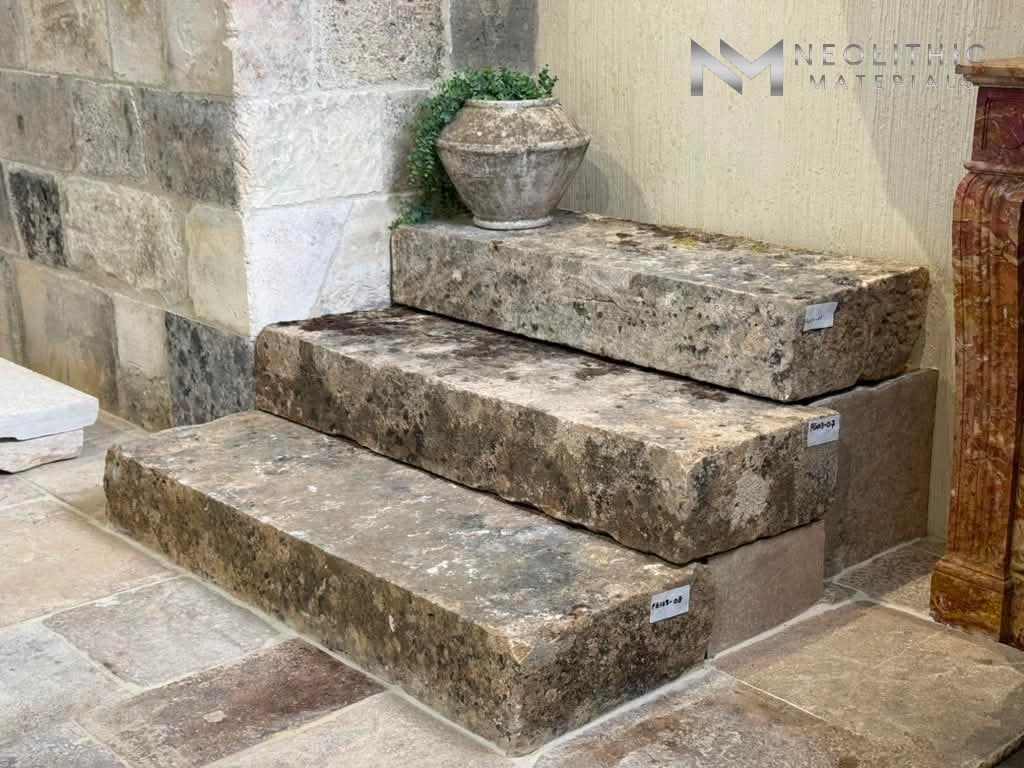 Tables and Slabs FG103-04, FG103-07 & FG103-08 indoor showroom installation shot showcasing three massive, rectangular antique stone blocks stacked into tiered steps, displaying a heavily weathered, moss-stained brown texture against a rustic stone brick wall and flooring.