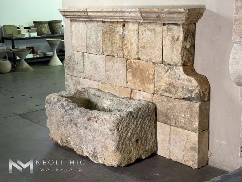 Troughs FG103-24.40x22x21 indoor showroom installation view capturing an ancient-style stone wall fountain featuring a high back constructed from rustic rectangular limestone blocks, complete with an architectural molding crown and a rugged, hand-chiseled rectangular basin trough at the base.