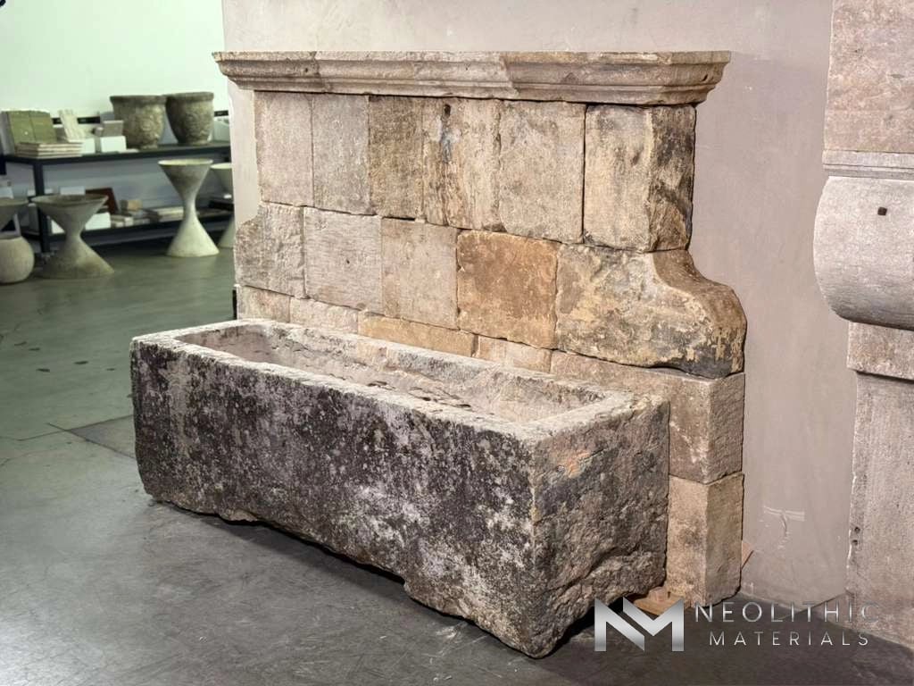 Fountains FG104-22.67x20x20 side-angle showroom display of an antique limestone wall fountain featuring a multi-block stone back panel with carved trim and a long, deeply weathered rectangular trough basin with a dark, aged patina.
