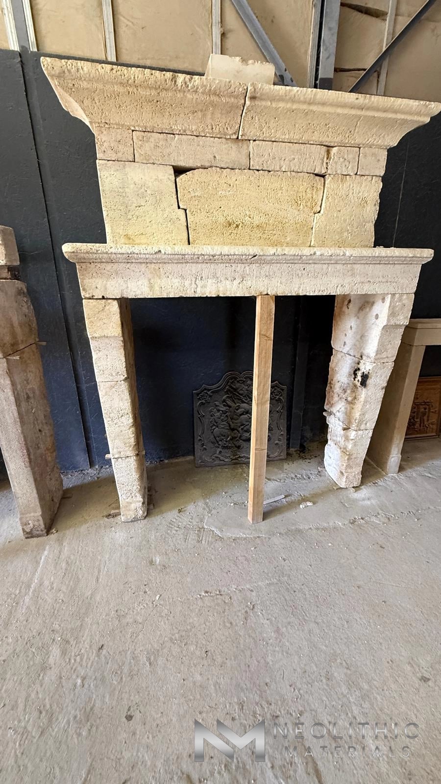 Reclaimed Fireplace Mantel FG105-02.NM eye-level indoor warehouse photograph showcasing an antique hand-carved stone mantelpiece surround featuring a rustic, pockmarked limestone texture and classical molding details supported by a wooden center post against a dark wall.