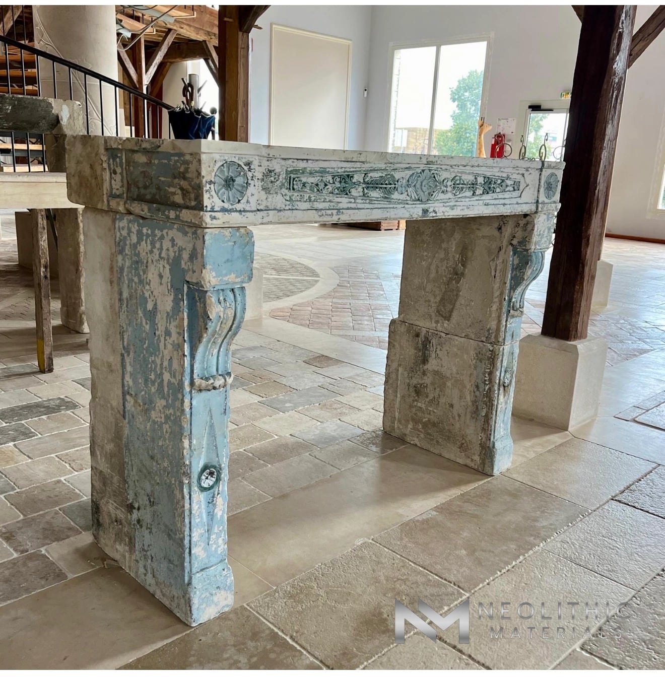 Reclaimed Fireplace Mantel FG105-03.N low-angle interior showroom photograph showcasing an ornate antique stone mantelpiece featuring distressed light blue and white painted finishes with classical scroll corbels standing on a tiled floor.