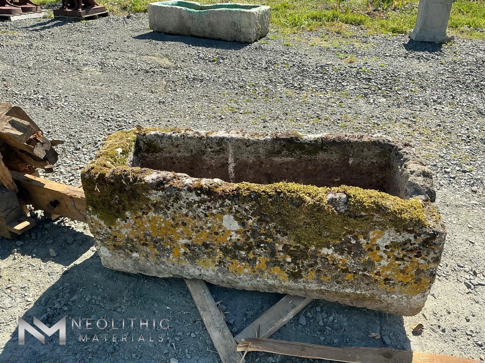 Trough FG105-05 high-angle outdoor photograph showcasing a long rectangular hand-carved stone basin covered in heavy green moss and yellow lichen growths resting on metal supports in a gravel salvage yard under bright sunlight.