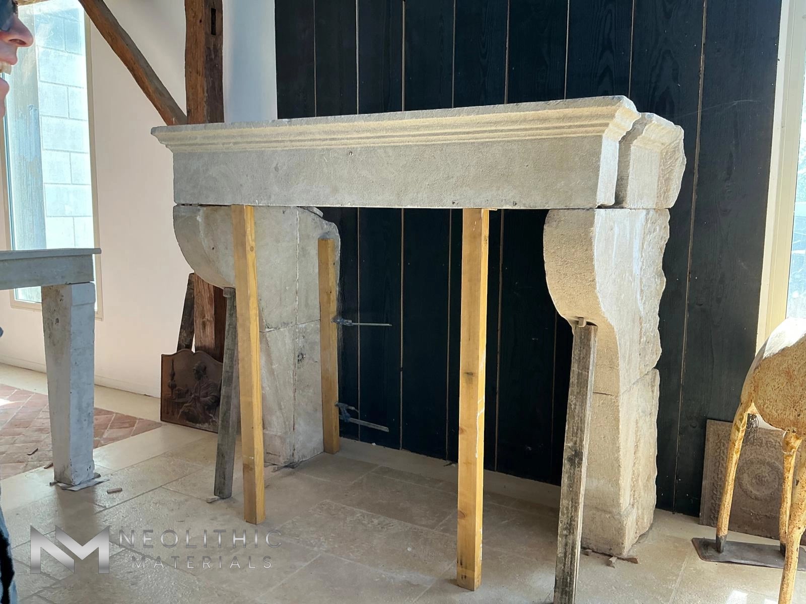 Reclaimed Fireplace Mantel FG105-18 angled interior showroom photograph highlighting an impressive, heavily weathered antique stone fireplace chimney piece featuring large carved scroll corbel legs and a deeply tiered molding mantle header supported temporarily by vertical wooden posts.