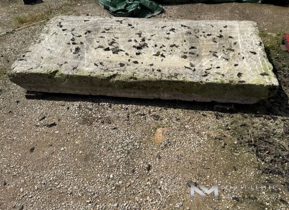 Tables and Slabs FK104-01 high-angle outdoor photograph looking down at a thick, rectangular hand-carved stone slab displaying a weathered pale grey face covered with scattered dry leaves and a mossy lower border on a sunlit gravel lot.