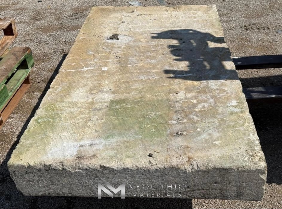 Tables and Slabs FK104-02 high-angle outdoor warehouse yard shot capturing a long, thick architectural limestone block casting a sharp worker's shadow across its distressed, sun-bleached grey surface face while resting elevated on dark support timbers.