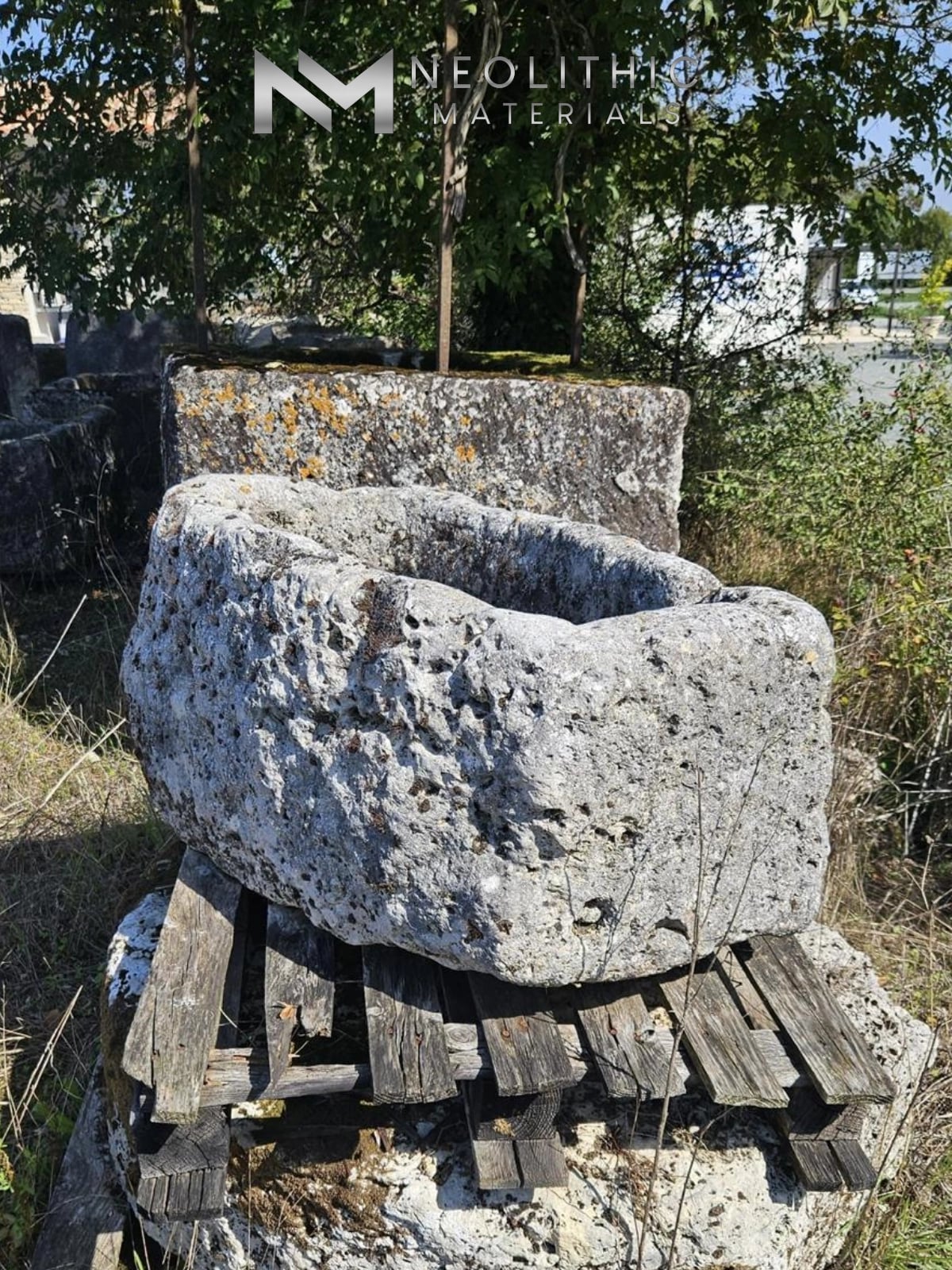 Trough FL101-07.ME close-up outdoor perspective view capturing a deeply pitted and weathered rectangular antique limestone basin resting on a sturdy wooden pallet with leafy green shrubbery growing in the sunny background.