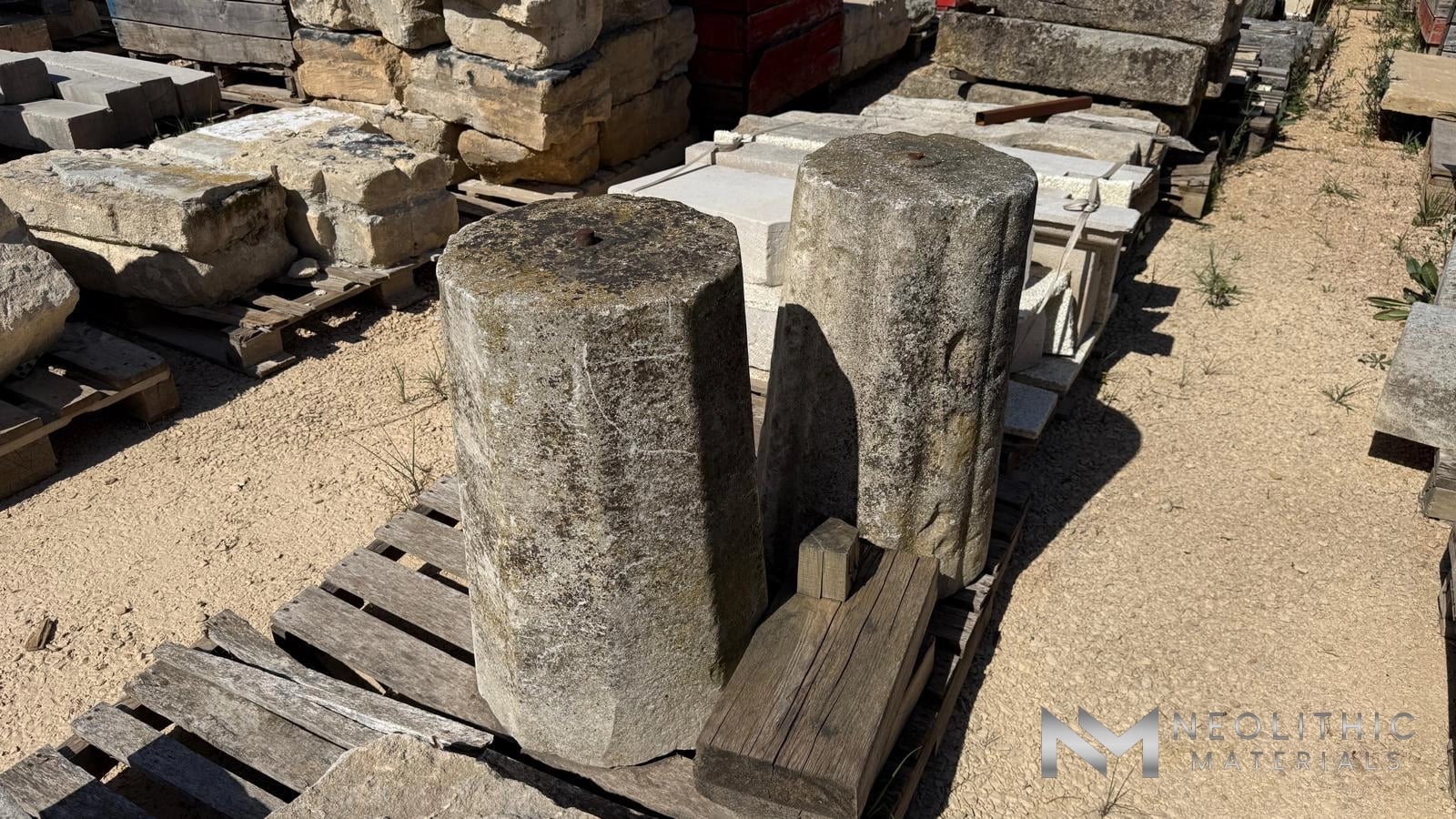 Miscellaneous FM101-01.MO & FM101-02.MO high-angle outdoor warehouse yard shot capturing two heavily weathered octagonal stone pillars standing upright on a wooden pallet next to a small carved masonry block on a sunny gravel lot.