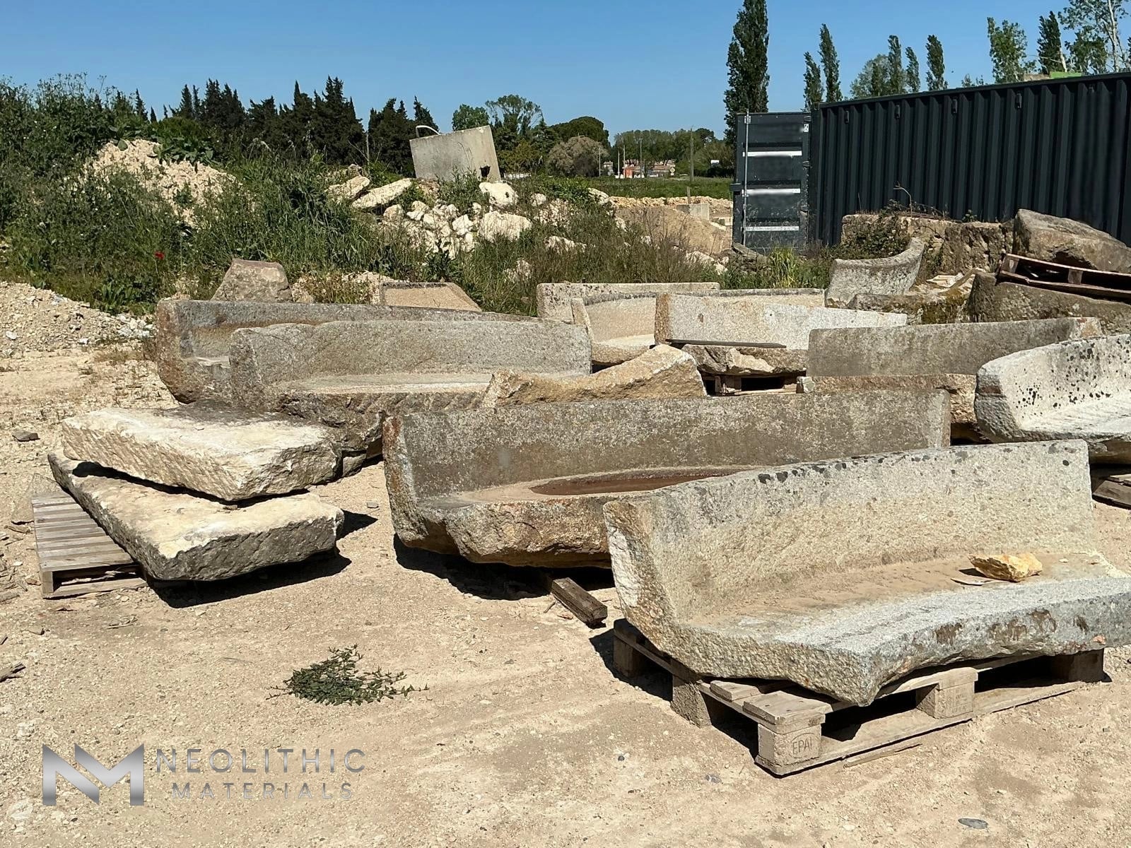 Bench FM101-03, 04, 05, 06 sunny mid-distance shot highlighting a collection of heavily weathered, crescent-shaped carved limestone benches arranged across a dry dirt lot with a dark shipping container and vegetation in the background.