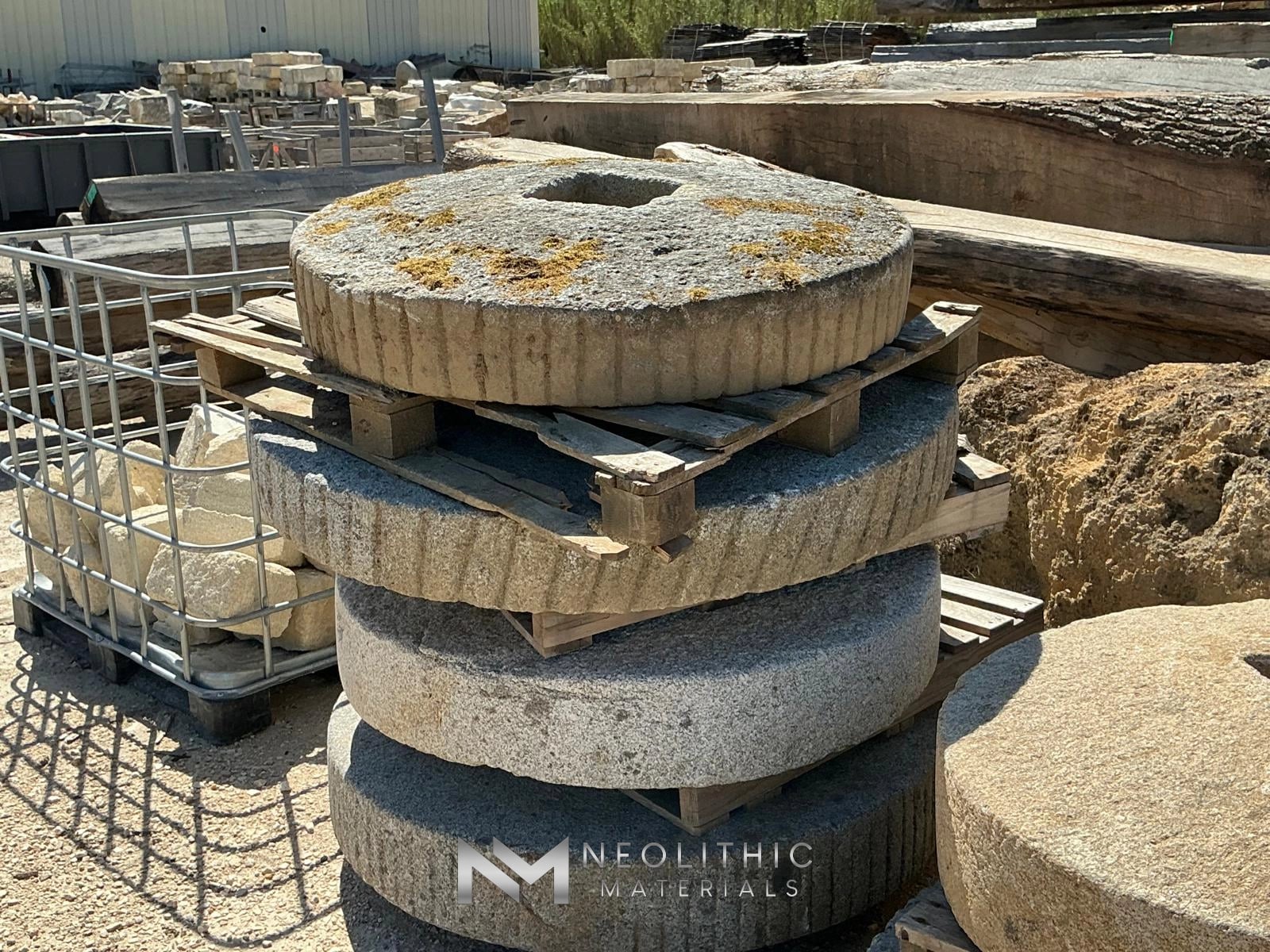 Trough FM101-07 & FM101-08 bright outdoor perspective shot focusing on a high stack of thick circular antique limestone wheels with square center cutouts stored on wooden pallets in an open masonry salvage yard.