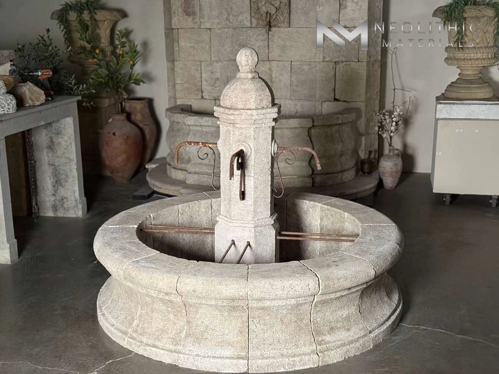 Fountains LA343-08.E.72x19x61 showroom display featuring a grand, ancient-style circular tier stone fountain with a tall central column pillar, showcasing a heavily weathered off-white limestone texture and ornate carved details.